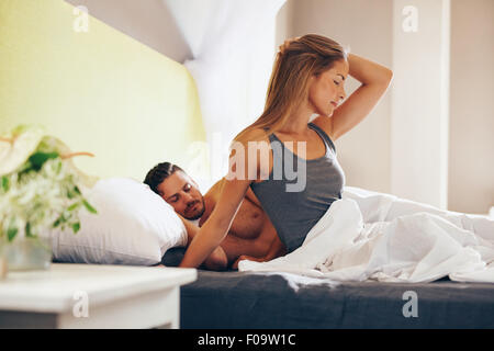 Portrait of attractive young woman waking up in morning with her husband sleeping behind her. Caucasian woman sitting on bed wit Stock Photo