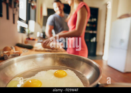 Fried eggs in a pan with couple standing in background are out of focus in domestic kitchen Stock Photo