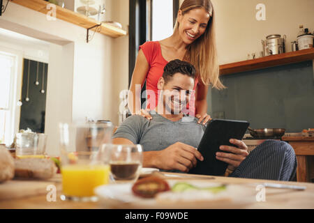 Young couple in kitchen looking at tablet pc smiling. Woman standing by her boyfriend sitting using digital tablet in morning. Stock Photo