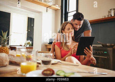 Young couple in kitchen looking at tablet pc. Man standing by his girlfriend sitting using digital tablet in morning. Stock Photo