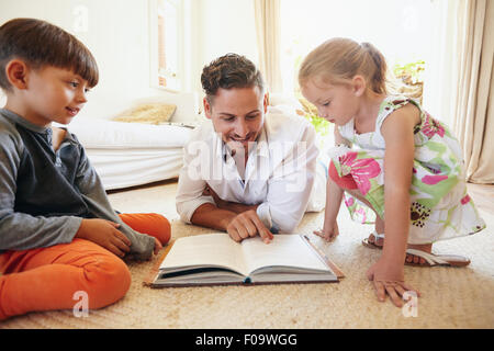 Family reading a book on the floor in the living room. Father reading stories to his son and daughter at home. Young man, little Stock Photo
