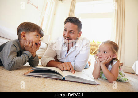 Shot of happy young family lying on the floor reading a book. Young man reading stories to his little son and daughter at home.