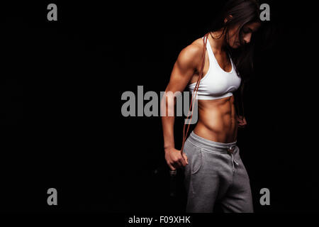 Image of female bodybuilder holding skipping rope looking down. Young fitness woman with muscular body posing on black backgroun Stock Photo