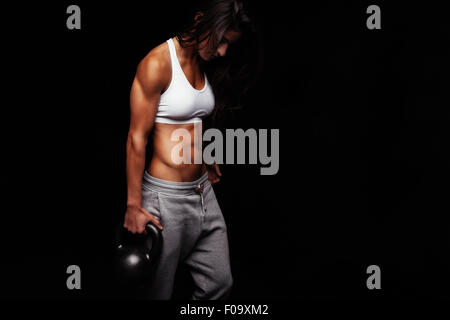 Young fit woman holding kettle bell exercising against black background. Muscular female doing crossfit exercise. Stock Photo