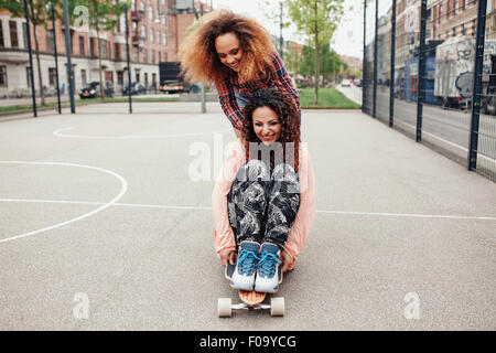 Woman pushing her friend on skateboard. Girls skating in basketball court. Young women having fun together outdoors with a longb