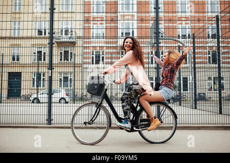 Portrait of two happy young women enjoying bike ride on city street. Female friends riding on one bicycle. Stock Photo