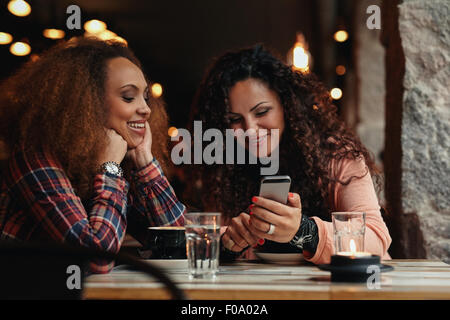 Two girls sitting at a table in cafe. One woman showing photos on her mobile phone to her friend. Stock Photo