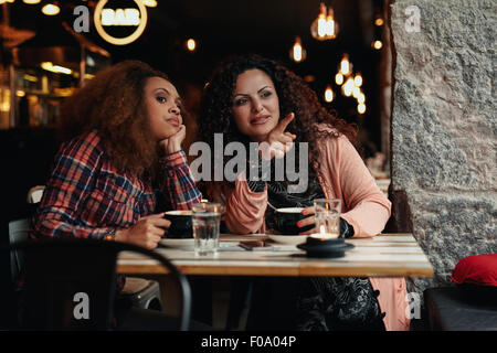 Two young women sitting at a restaurant looking away, with one pointing away showing something to her friend. Stock Photo