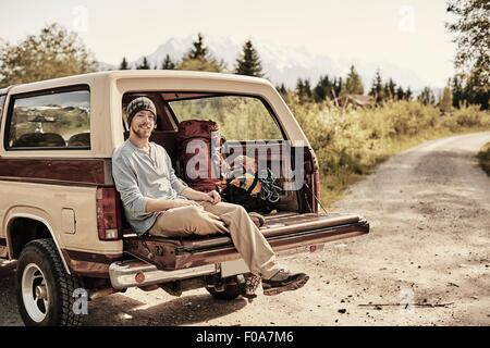Young man sitting on back of pickup truck, looking at camera, smiling Stock Photo