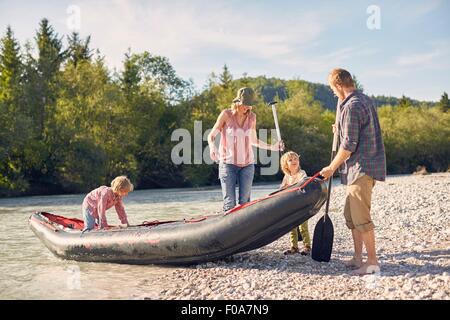 Family pulling dinghy onto riverbank, holding paddle boards Stock Photo