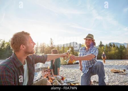 Adult men sitting around campfire making a toast with beer bottles Stock Photo
