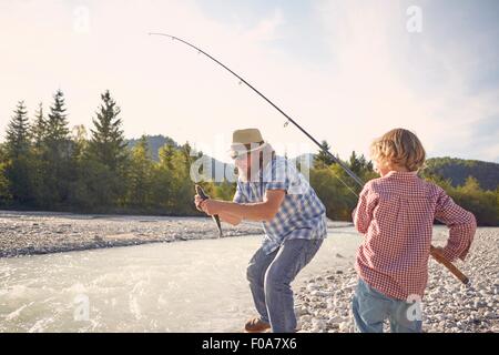 Mid adult man and boy next to river using fishing rod to catch fish Stock Photo