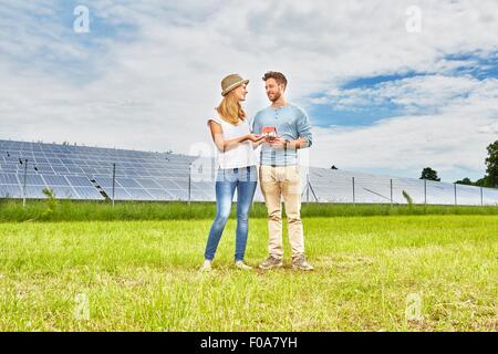 Young couple standing in field, holding small model of house, next to solar farm Stock Photo