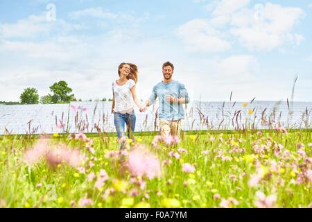 Young couple holding hands, running through field, next to solar farm