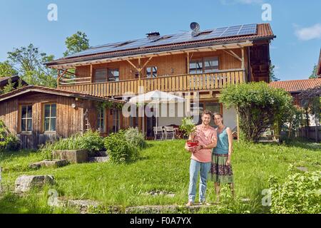 Portrait of young couple, holding pot plant, standing in front of house with solar panelled roof Stock Photo