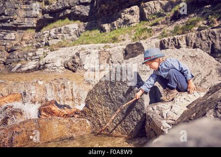 Young boy, sitting on rock, prodding mud with stick Stock Photo