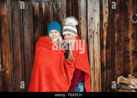 Two young female friends wrapped in red blanket outside wooden cabin
