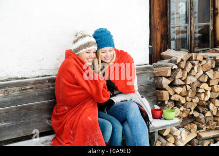 Two young female friends wrapped in red blanket sitting  outside wooden cabin