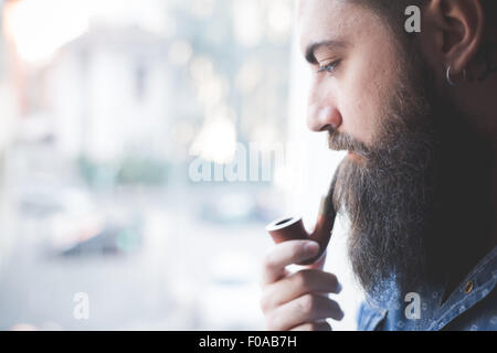 Young bearded man smoking pipe by window Stock Photo