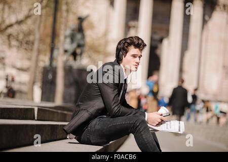 Young city businessman with newspaper sitting on bench listening to headphones Stock Photo