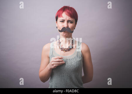 Studio portrait of confused young woman holding up mustache in front of face Stock Photo