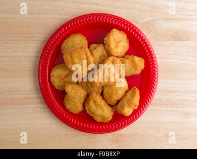 Top view of a serving of chicken nuggets on a red plate on a wood table top illuminated with window light. Stock Photo