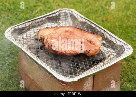 A bacon steak being cooked on a disposable barbecue outdoors Stock Photo