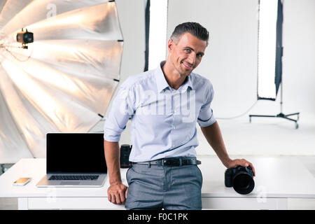 Portrait of a smiling confident photographer leaning on the table in studio and looking at camera Stock Photo