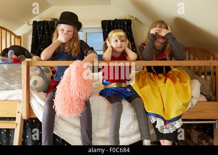 Three sisters, sitting on bed,  wearing fancy dress costumes, covering mouth, ears and eyes Stock Photo