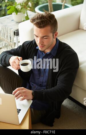 Portrait of man sitting on floor at home looking at laptop and drinking cup of coffee Stock Photo