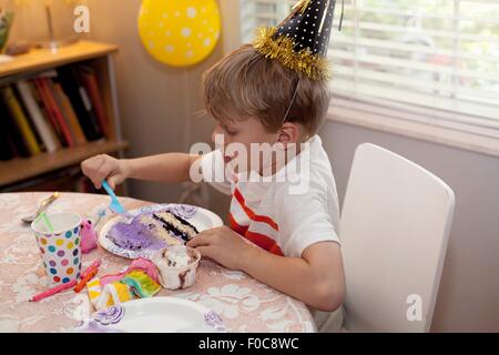 Boy in party hat sitting at table eating birthday cake Stock Photo