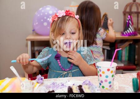 Portrait of girl sitting at birthday party table eating purple cake with purple tongue Stock Photo