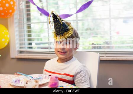 Boy in party hat sitting at table eating birthday cake pulling funny face Stock Photo