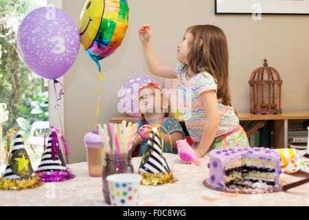 Two girls sitting at birthday party table with cake playing with balloons Stock Photo