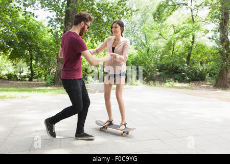 Young man assisting woman in skateboarding on footpath at park Stock Photo