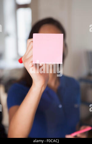 Businesswoman writing on adhesive note stuck on glass wall Stock Photo