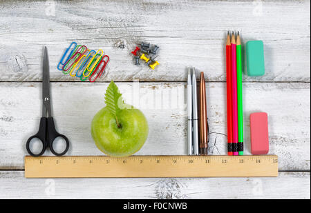 Green apple, pens, pencils, erasers, paper clips, tacks and scissors lined up with ruler on top of white desktop. Stock Photo