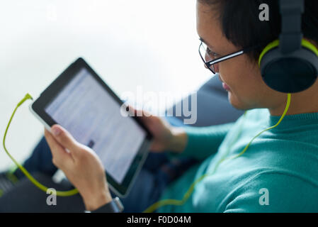 Chinese man relaxes on sofa and watches podcast on tablet pc, listening with green big earphones. Copy space Stock Photo
