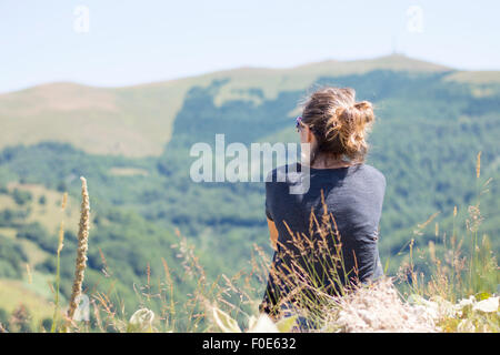 Young woman with backpack sitting on cliff's edge and looking to mountains and clear sky. Summer hiking trip adventure Stock Photo