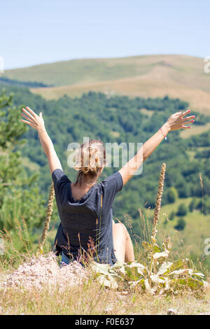 Young woman with backpack sitting on cliff's edge and looking to mountains and sky with raised hands. Hiking trip joy Stock Photo