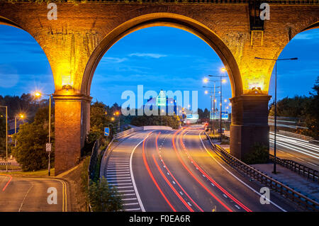 Railway viaduct crossing over M60 motorway near Manchester, UK. Tail lights of cars leaving trail passing under the bridge. Stock Photo