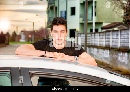 Serious young man sitting on his car's door, resting on the roof and looking at camera Stock Photo