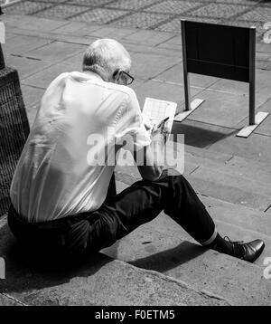 Photo of an elderly man sitting on steps and doing a crossword or sudoku puzzle Stock Photo