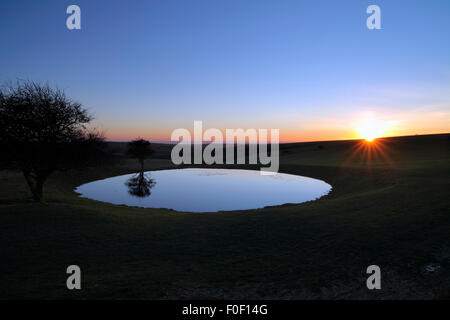 The sun setting behind a dew pond on the South Downs, near Ditchling Beacon, East Sussex. Stock Photo