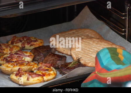 Hand in the oven-glove taken out from the oven oven-tray with steaks, potato with cheese and bacon and Greek pitas. Stock Photo