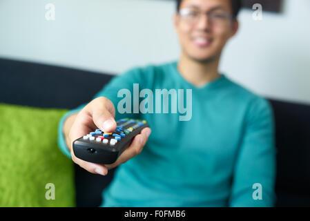Asian young adult man watching TV and changing channel with remote control. Narrow focus on buttons and hand in foreground Stock Photo