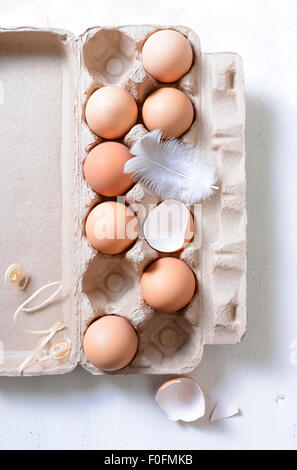 Farm fresh eggs in egg carton, with chicken feather and broken shells on white vintage wood table. Stock Photo