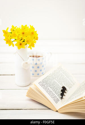 small bunch of daffodils on white table, with mug in the background and open book with woodpecker feather Stock Photo