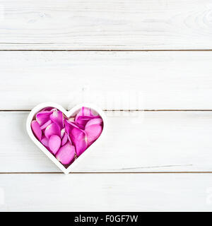 a white heart shaped bowl filled with pink rose petals, on a whitewashed wooden background Stock Photo