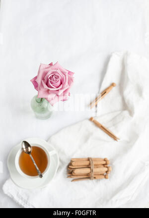 a single pink rose in focus, with blurred background of white fabric, teacup with teaspoon, and dolly clothes pegs Stock Photo